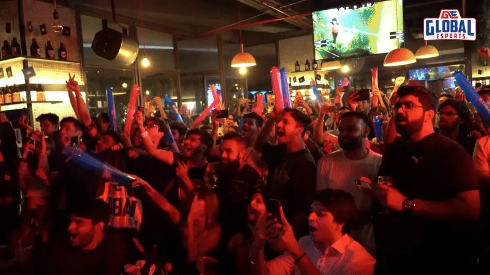 A crowd of fans cheering in a bar under a big screen with the GE logo